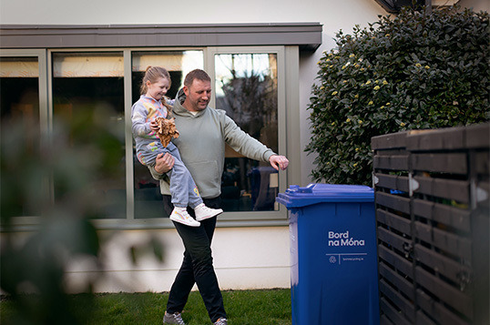 Father carrying daughter over to the blue Bord na Móna wheelie bin in their back garden to put paper waste in it.
